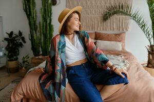 romántico mujer con sincero sonrisa sentado en cama, disfrutando soleado Mañana en su elegante plano en boho estilo. foto