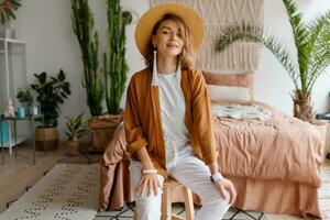 romántico mujer con sincero sonrisa sentado en cama, disfrutando soleado Mañana en su elegante plano en boho estilo. foto