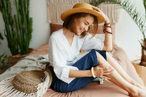 Romantic woman with candid smile sitting on bed, enjoying sunny morning in her stylish flat in boho style. photo