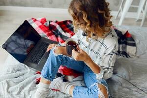 Stylish smart cheerful  woman in  eyeglasses using laptop , lying on cozy bed, drinking coffee. photo