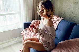 Close up indoor portrait of graceful blonde woman enjoying smell of cappuccino  , dreaming and looking into the window.  Wearing pink knitted sweater. photo