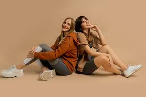 Two laughing sportive  women chilling after training,  sitting on floor in studio on beige background. photo