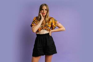 Studio fashion photo of pretty blond woman in stylish top and  linen shorts posing on purple background.