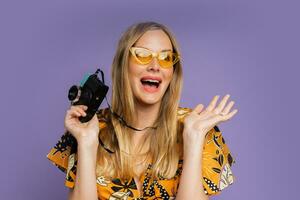 Pretty blond woman in stylish summer outfit holding camera and posing on purple background in studio. Travel and summer fashion concept. photo