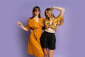 Two playful stylish women posing in studio over purple background. Friends dancing and having fun. photo