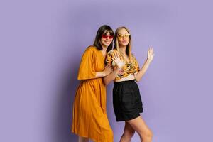 Two playful stylish women posing in studio over purple background. Friends dancing and having fun. photo
