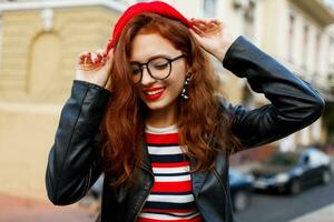 elegante joven jengibre mujer en rojo boina y lentes posando en el calle. foto