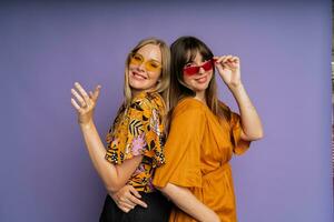 cerca arriba retrato de dos elegante mujer en Gafas de sol y de moda verano ropa posando en púrpura fundamento en estudio. foto