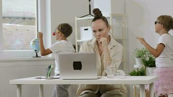 une femme séance à une bureau avec une portable avec les enfants en jouant autour video