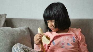 liten flicka Sammanträde på soffa äter en banan. video