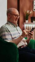 Vertical Video Retired traveller browses smartphone app in lounge area at five star holiday retreat, using online network to navigate on webpage. Senior client at hotel relaxing in lobby.