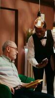 vertikal video pensionerad gäst betalande för kaffe använder sig av kreditera kort på hotell, beställning dryck från bar och pos terminal betalning. manlig concierge tjänande äldre resande med förfriskning medan väntar.