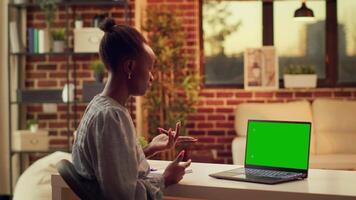 Woman waves at greenscreen on pc, talking to someone on videocall and working on freelancing career. Person using laptop with blank mockup display, attend internet meeting at sunset. video