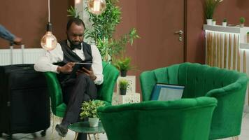 African american hotel staff wearing uniform in lobby using digital tablet to help manage online room bookings. Bellhop holding electronic device entering details for upcoming reservations. video