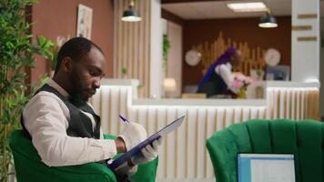 In lobby, employee in tuxedo reviews inventory of bookings. Hotel administrator working as lodging manager and dealing with room reservations on papers. Bellboy handles list of guests. video