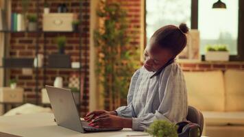 Teleworker takes phone call while working on blog articles at home, talking to editor to solve some job related problems. African american person using telephone line for development. video