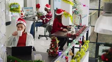 Zoom in portrait shot of happy asian employee working on christmas season in decorated office. Smiling worker solving tasks at desk in diverse xmas ornate workplace next to BIPOC colleagues video