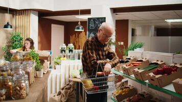 ritratto di sorridente anziano cliente guardare per dispensa graffette su zero rifiuto supermercato scaffali. contento anziano uomo acquisto biologico cibo nel riutilizzabile barattoli a Locale drogheria memorizzare video