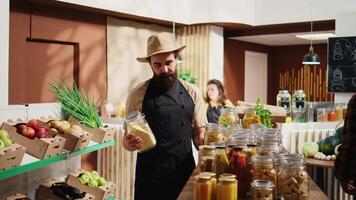 Portrait of smiling vendor adding pantry staples on sustainable zero waste supermarket shelves. Happy seller restocking local neighborhood grocery store with organic food in reusable jars, close up video