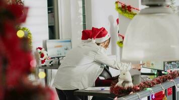 afrikansk amerikan anställd arbetssätt i jul utsmyckad kontor begärande hjälp från kollega. företag personal ser över finansiell siffror på xmas smycka arbetsplats dator skärm video