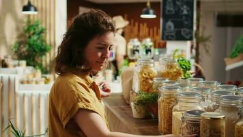 Woman using smartphone in zero waste supermarket to check what ingredients she needs for healthy recipe she plans to cook. Customer in local grocery shop uses phone while shopping for organic veggies video