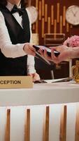 Vertical Video Senior man makes nfc payment with mobile phone at pos terminal, paying for room at five star hotel. Person arriving at reception desk and doing check in for vacation. Close up. Handheld