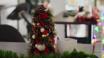 Close up shot on miniature Christmas pine tree decoration placed in empty startup office, with blurry background of festive workplace filled with xmas lights, garlands and wreaths video