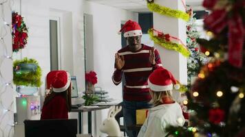African american employee wearing Christmas hat entertaining coworkers by playing pretending game during break. Colleague having fun doing pantomime during Secret Santa party video