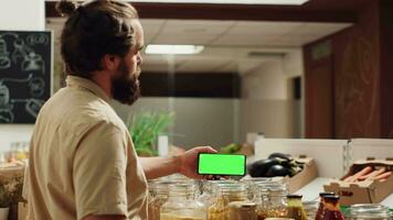 Man using green screen landscape mode smartphone in eco friendly supermarket. Client in local grocery shop uses chroma key phone while shopping for organic veggies, panning shot video