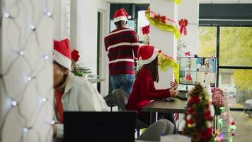 Asian staff member in videocall with teammates during Christmas season in festive decorated office. Employee in teleconference meeting with team in busy xmas ornate workplace video