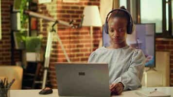 africano americano donna multitasking, finitura compiti Più veloce Se lei ascolta per musica a casa stazione di lavoro durante tramonto ora. libero professionista edificio in linea carriera e godendo Audio melodie. video