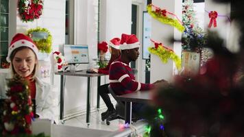 retrato de sonriente africano americano empresario trabajando en Navidad temporada en decorado oficina. empleado resolviendo Tareas a escritorio en diverso lugar de trabajo siguiente a compañeros de trabajo durante invierno festivo hora video