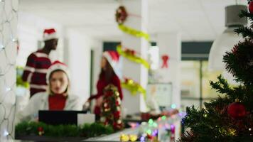 Focused employee in Christmas decorated office working on overdue project while carefree coworkers have fun together looking at decorations. Stressed worker in xmas adorn workspace video