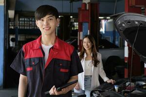 retrato de joven asiático masculino automotor mecánico trabajador con hembra clientes ev coche a mantenimiento cochera, contento sonrisas, profesional Servicio estación, cheque y reparar en auto transporte industria. foto