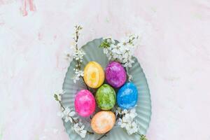 Colorful Easter eggs on plate and spring white cherry blossom flowers on pink background. Easter celebration concept. Top view, flat lay, copy space photo