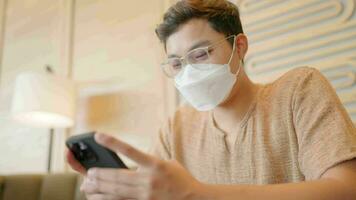 Asian man wearing a mask is typing a message on a smartphone and looking at the window. video