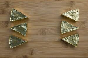 Pieces of processed cream cheese spread on wooden cutting board. Family portion of six triangles in gold foil. Minimalist background. Closeup, top view, copy space photo