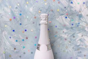 White champagne party bottle and colorul metallic stars confetti on blue background. Flat lay, copy space. New year and Christmas celebration concept photo