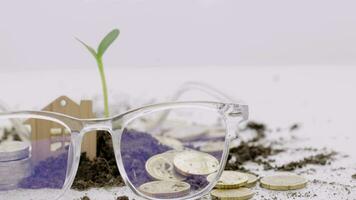 a house, glasses, coins and a plant with soil on the table, financial growth concept video