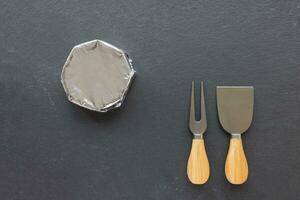 French round camembert cheese in wrapping foil paper and cheese knives on black slate plate. Closeup, top view, copy space photo