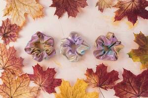 Pink metallic holographic scrunchies and autumn red and yellow maple leaves on pastel background. Trendy accessories from 80s and 90s. Flat lay, top view. DIY, hairstyle, lifestyle, outfit ideas for photo