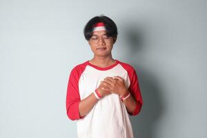 Portrait of attractive Asian man in t-shirt with red and white ribbon on head, placing hands on chest, feeling peaceful. Isolated image on gray background photo
