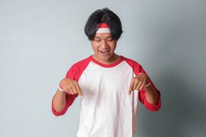 Portrait of attractive Asian man in t-shirt with red and white ribbon on head, pointing at something down with finger. Isolated image on gray background photo