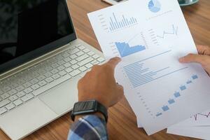 Close up of male freelance worker hand working on laptop and pointing with finger on analysis data with business graph and chart of information diagram on office desk in the morning photo
