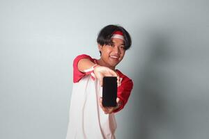 Portrait of attractive Asian man in t-shirt with red and white ribbon on head, holding and showing blank screen of mobile phone for mock-up. Isolated image on gray background photo