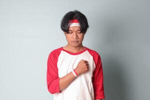 Portrait of attractive Asian man in t-shirt with red and white ribbon on head, placing his hand on chest, giving respect and salute hand gesture. Isolated image on gray background photo