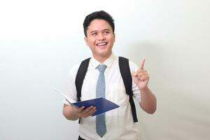 Indonesian senior high school student wearing white shirt uniform with gray tie pointing at empty space while holding an opened note book. Isolated image on white background photo