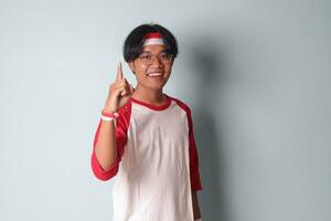 Portrait of attractive Asian man in t-shirt with red and white ribbon on head, presenting index finger and reminding of something to do. Isolated image on gray background photo