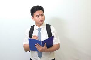 Indonesian senior high school student wearing white shirt uniform with gray tie writing on note book using pen and thinking about an idea. Isolated image on white background photo