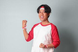 Portrait of attractive Asian man in t-shirt with red and white ribbon on head, raising his fist, celebrating Indonesia's independence day. Isolated image on gray background photo